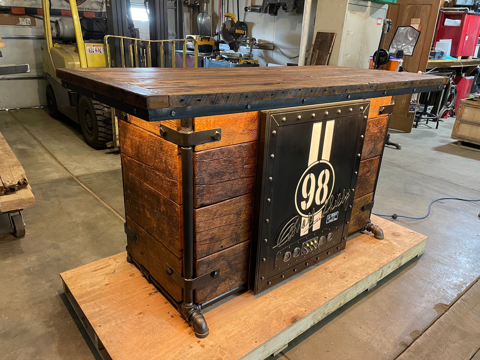 A large industrial-style bar made of barn wood with a hidden fridge and a Carroll Shelby door front, featuring LED lighting and stewert Warner gauges.